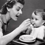 A vintage black-and-white photograph of a mother spoon-feeding a baby in a high chair
