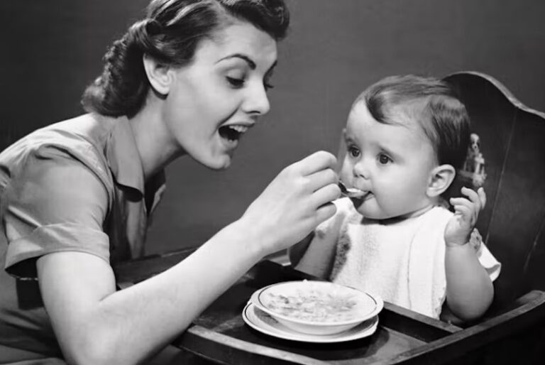 A vintage black-and-white photograph of a mother spoon-feeding a baby in a high chair