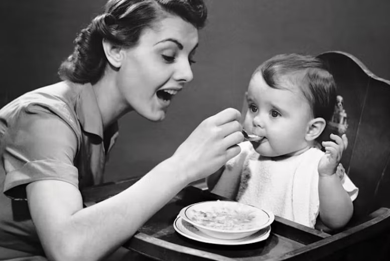 A vintage black-and-white photograph of a mother spoon-feeding a baby in a high chair