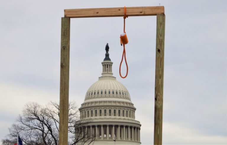 A wooden gallows with a hanging noose, erected by the crowd outside the United States Capitol, framing the Capitol dome