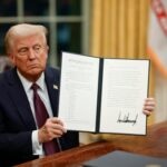 Donald Trump in the Oval Office holding up the signed Proclamation 10887, the blanket January 6 pardon, on the first night of his second term
