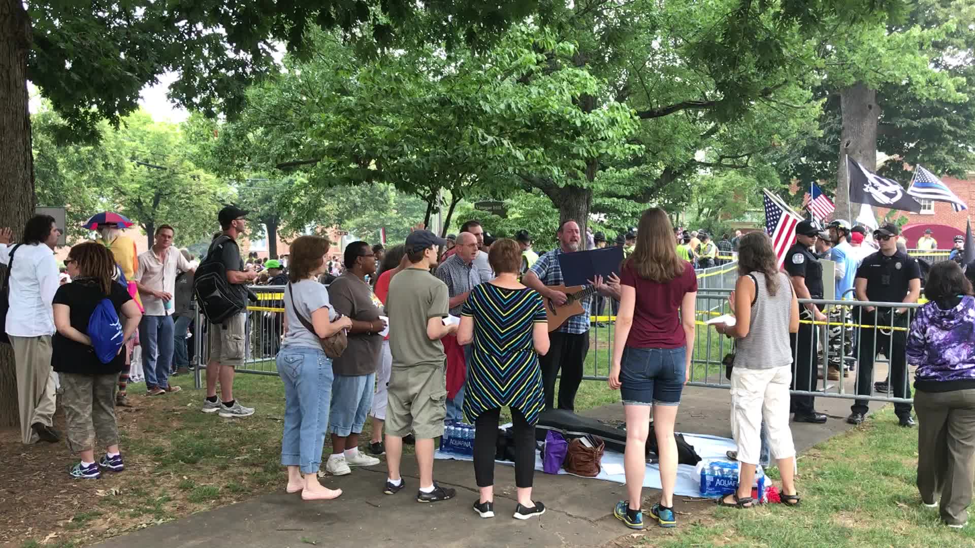 Crowds line metal barricades under the trees at the edge of Emancipation Park in Charlottesville, August 12, 2017; nationalist flags are visible among the rally-goers in the background, and cases of water are staged along the fence.